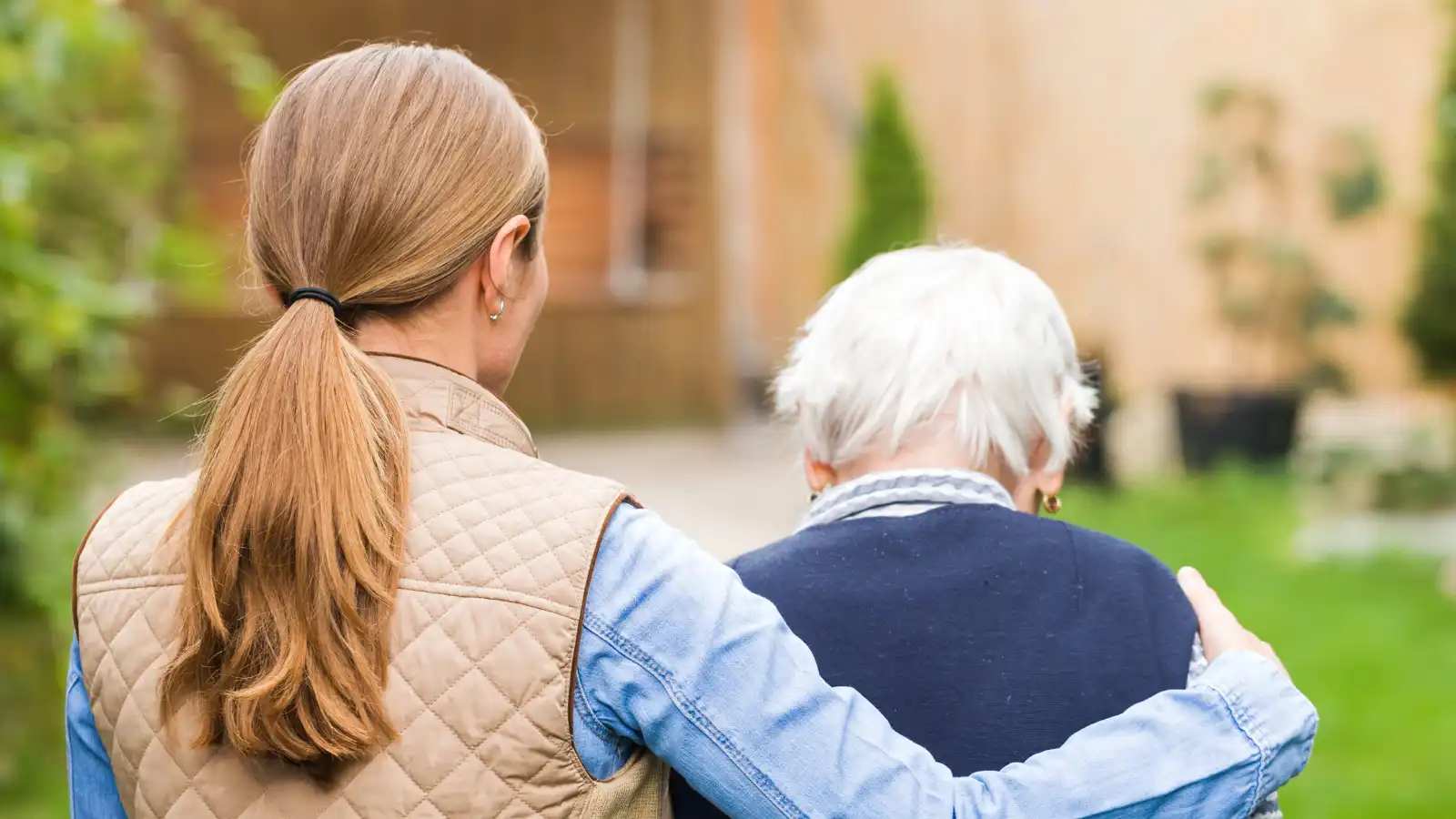 Younger woman supporting an elderly woman outdoors, both seen from behind